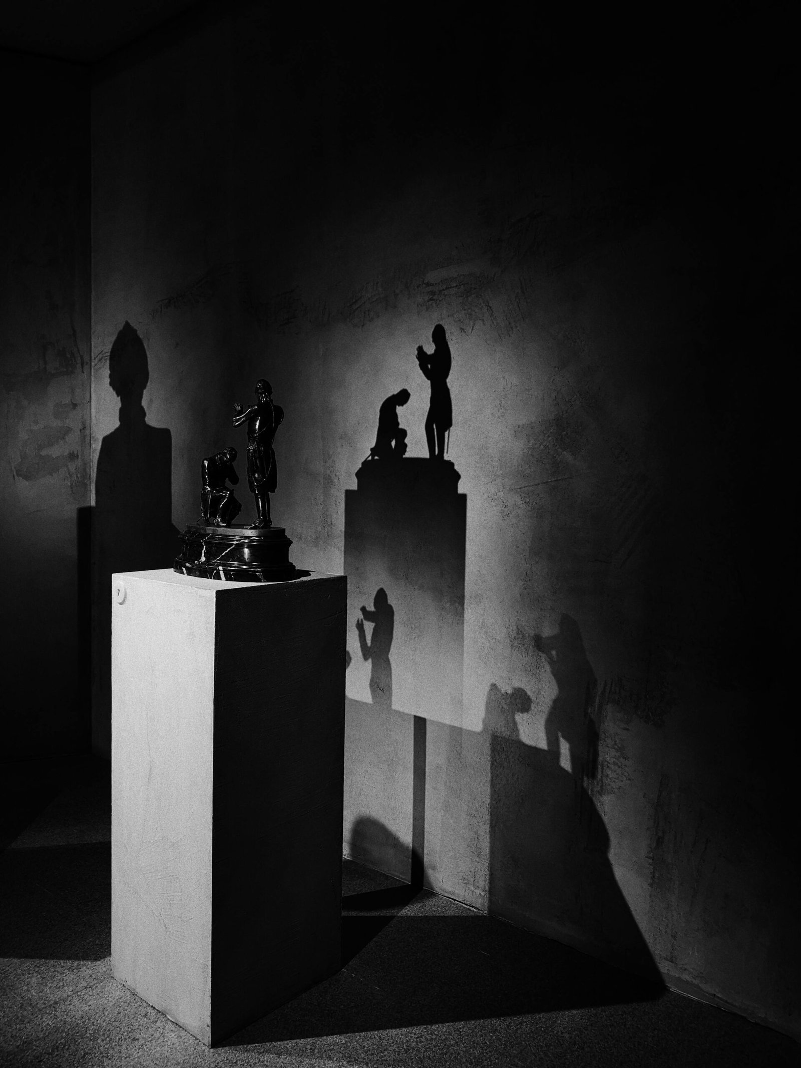 Black and white image capturing shadows of sculptures on a gallery wall with strong contrast.